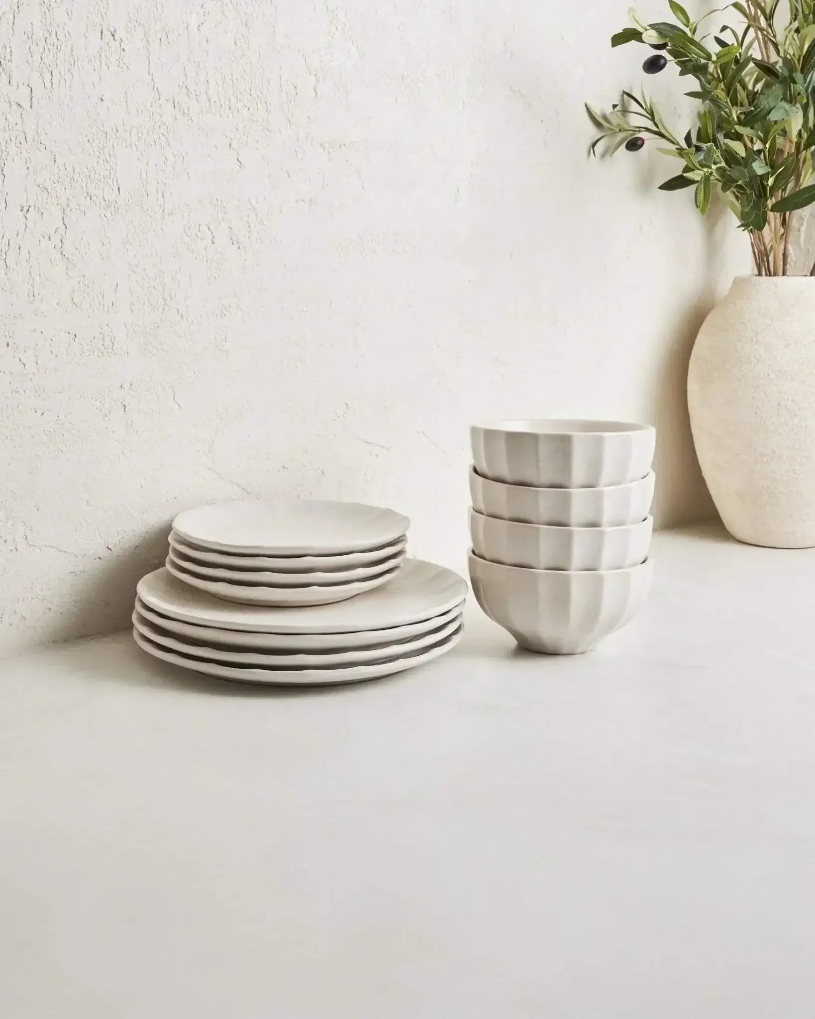 Stacked white ceramic plates and bowls on a neutral countertop with a decorative vase