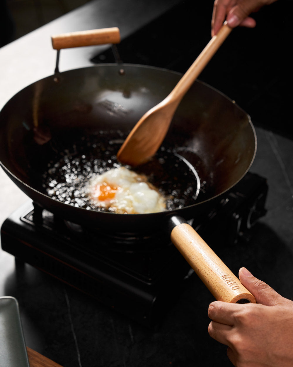 Cooking a fried egg in a MAKO wok on a stovetop with a wooden spatula