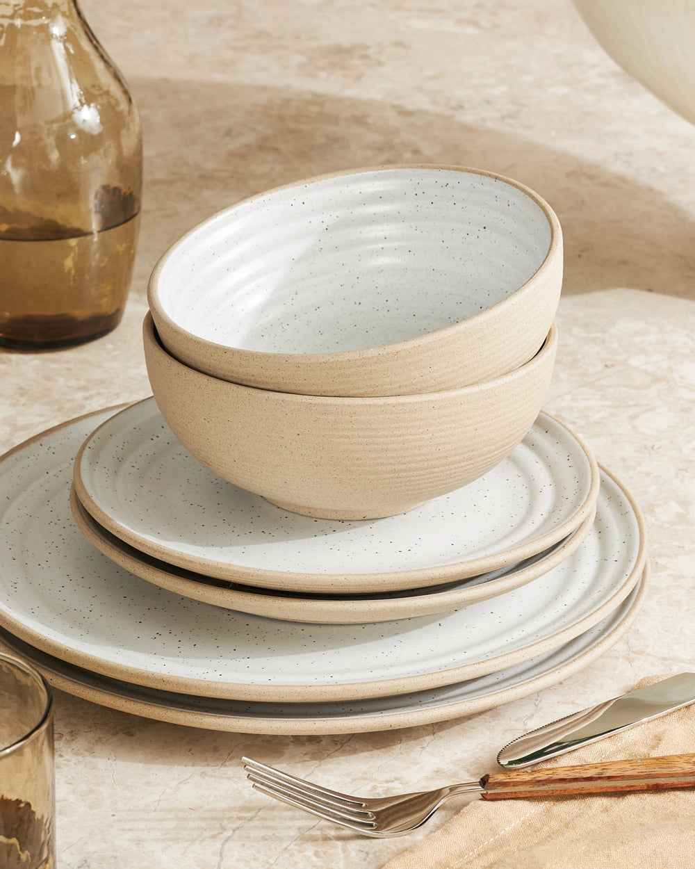 Set of four speckled ceramic cereal bowls and plates on a neutral stone table.
