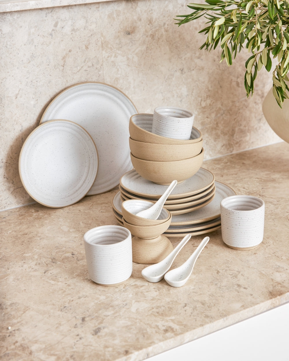 Neutral-toned ceramic dinnerware set with plates, bowls, cups, and spoons on a stone counter
