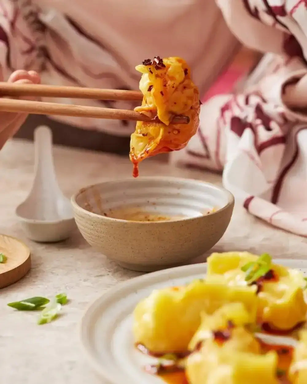 Chopsticks holding spicy yellow dumpling above dipping bowl with sauce, plate of dumplings on table