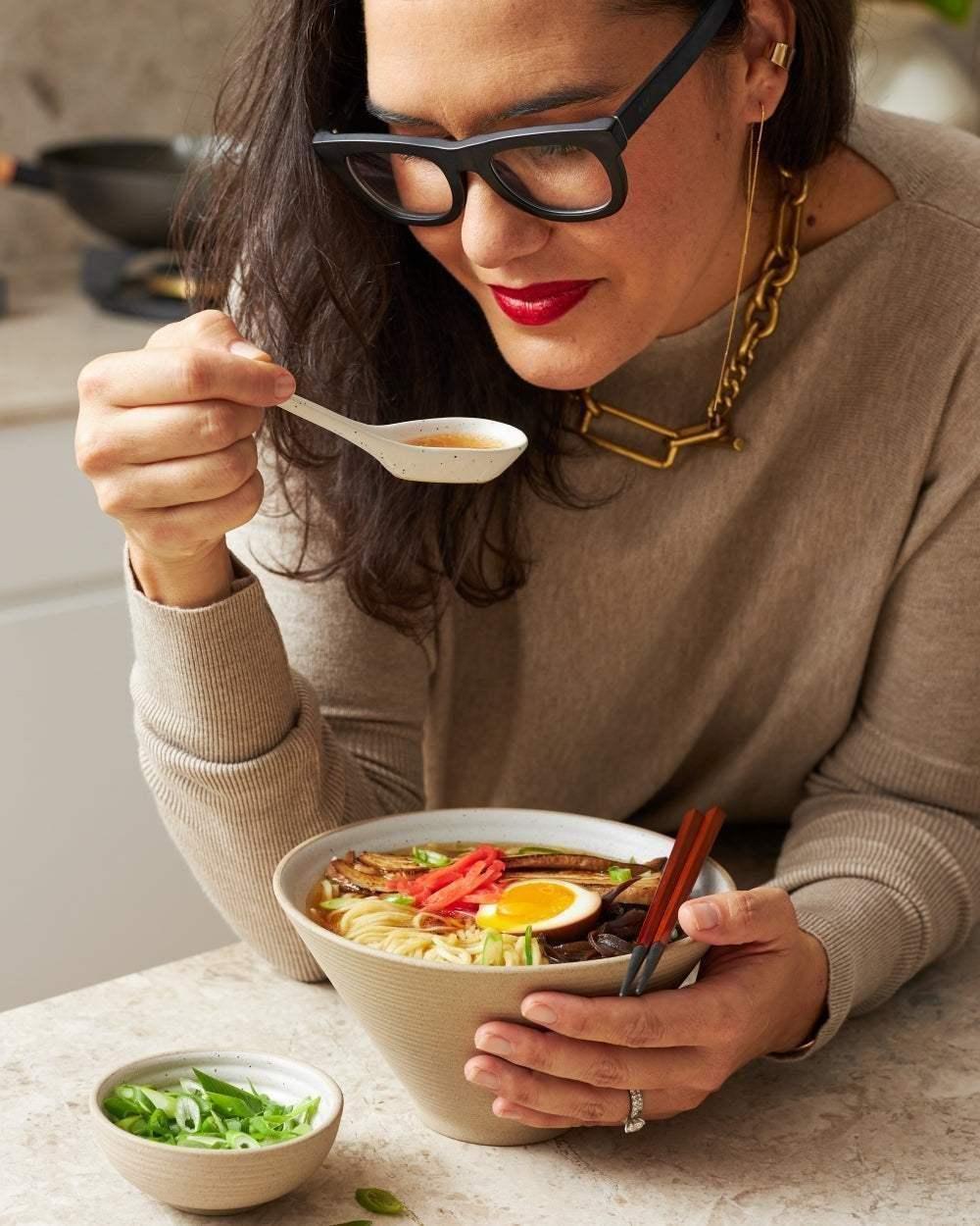 Woman enjoying noodle soup with an egg in a textured ceramic bowl using chopsticks and spoon