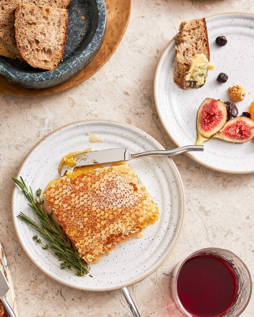 Speckled ceramic side plates with honeycomb, figs, bread, and a glass of red wine on a table.