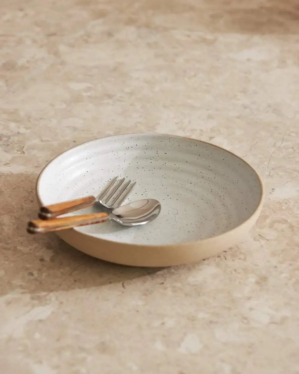 Asoke wide ceramic bowl with speckled white glaze, set on stone countertop with fork and spoon with wooden handles