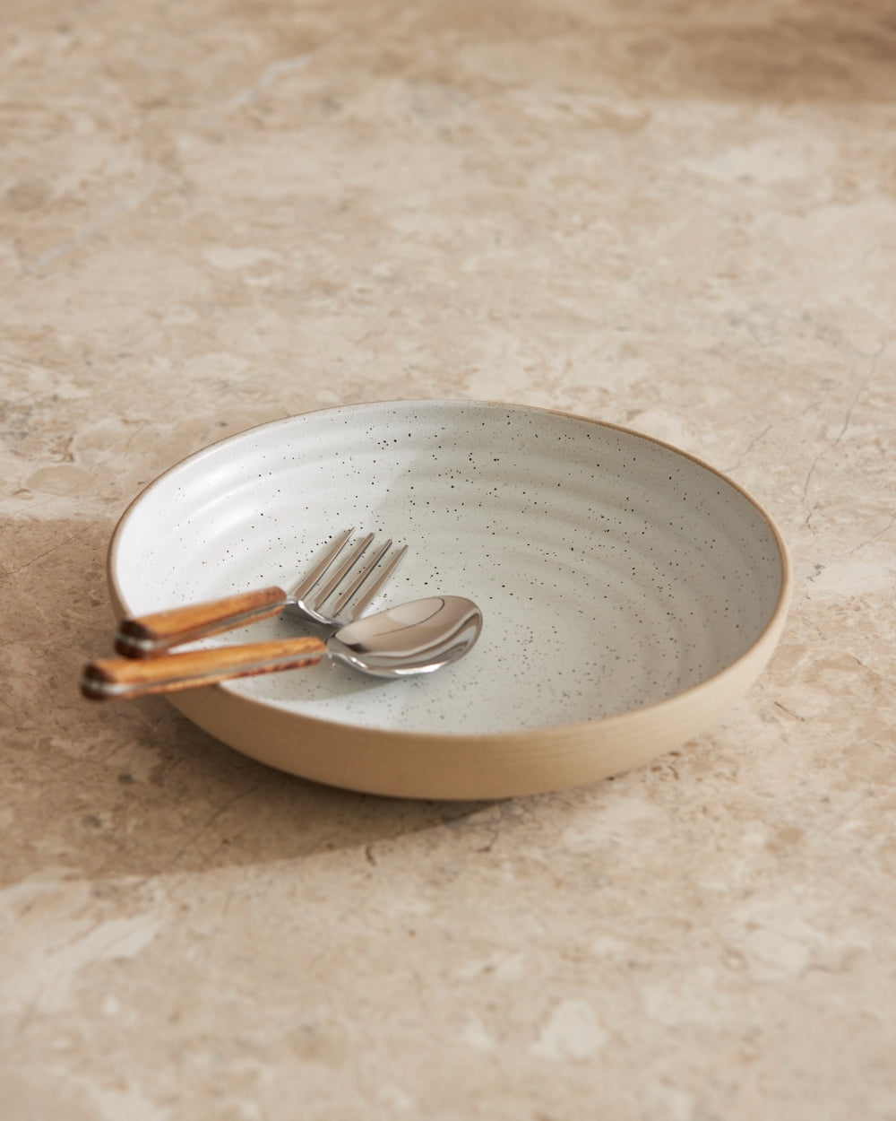 Wide ceramic bowl with speckled glaze, spoon and fork with wooden handles on stone countertop