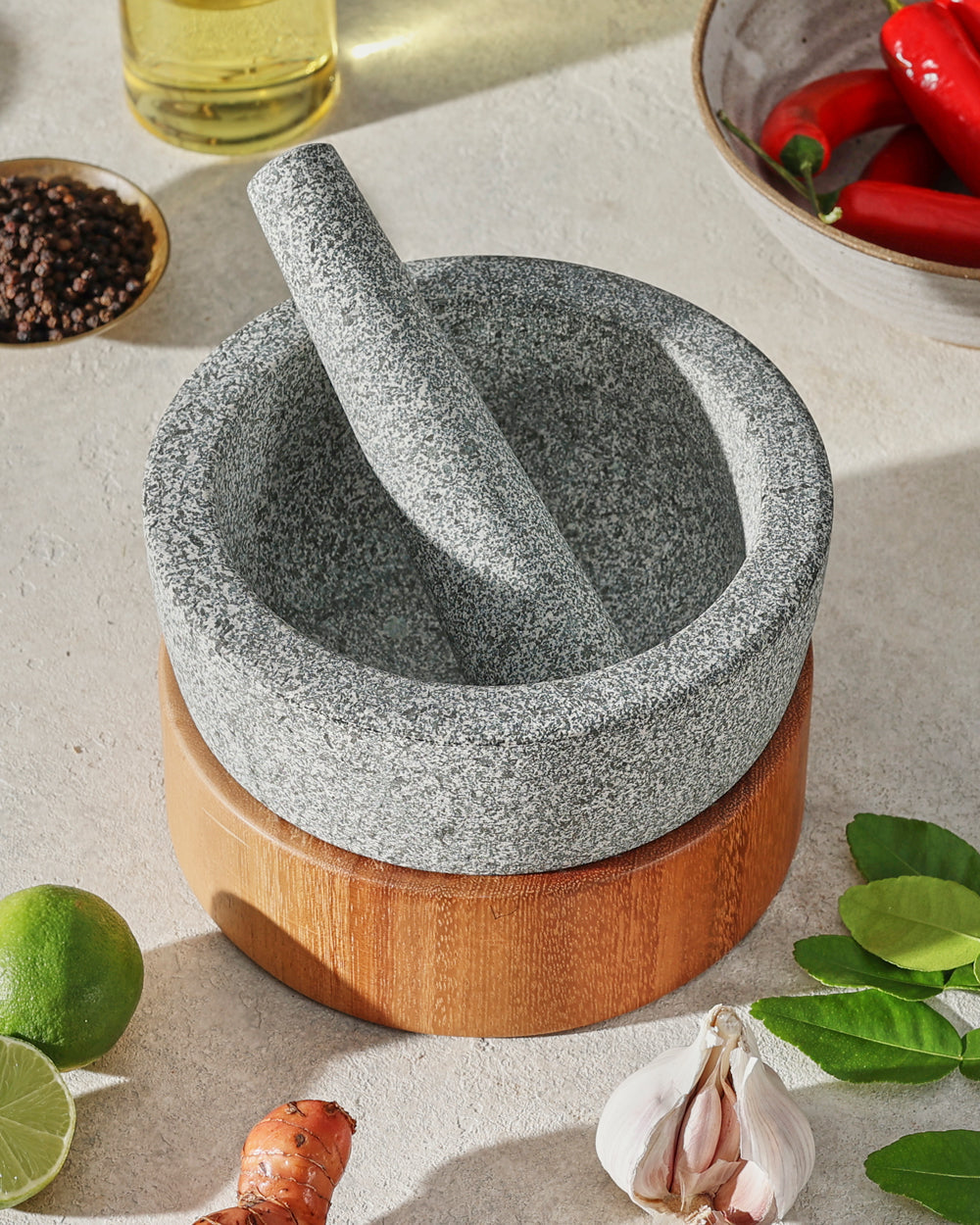 Gray stone mortar and pestle with wooden base surrounded by fresh herbs, lime, garlic, and chili.