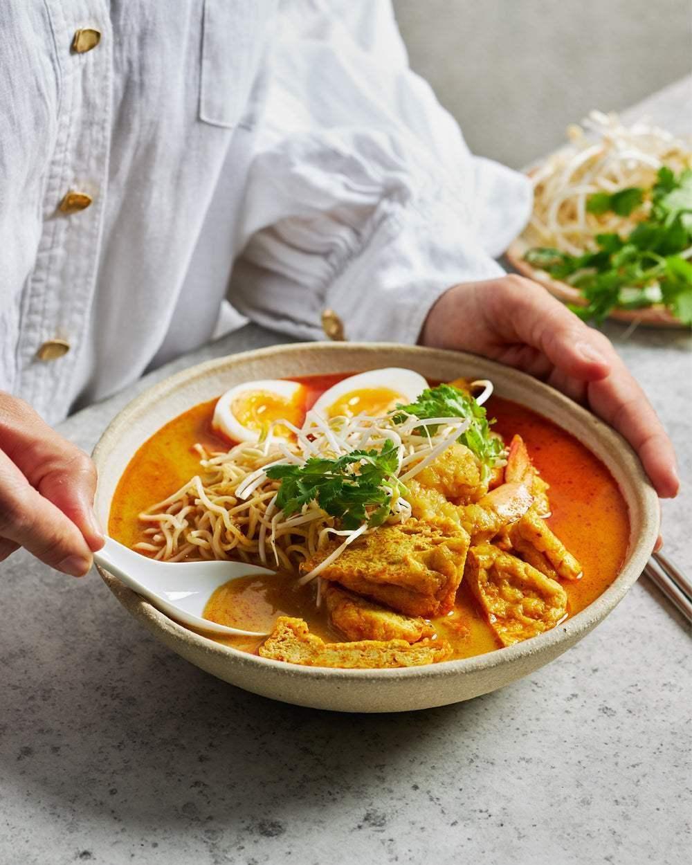 Colorful bowl of noodle soup with egg, tofu, bean sprouts, and fresh herbs on a grey surface