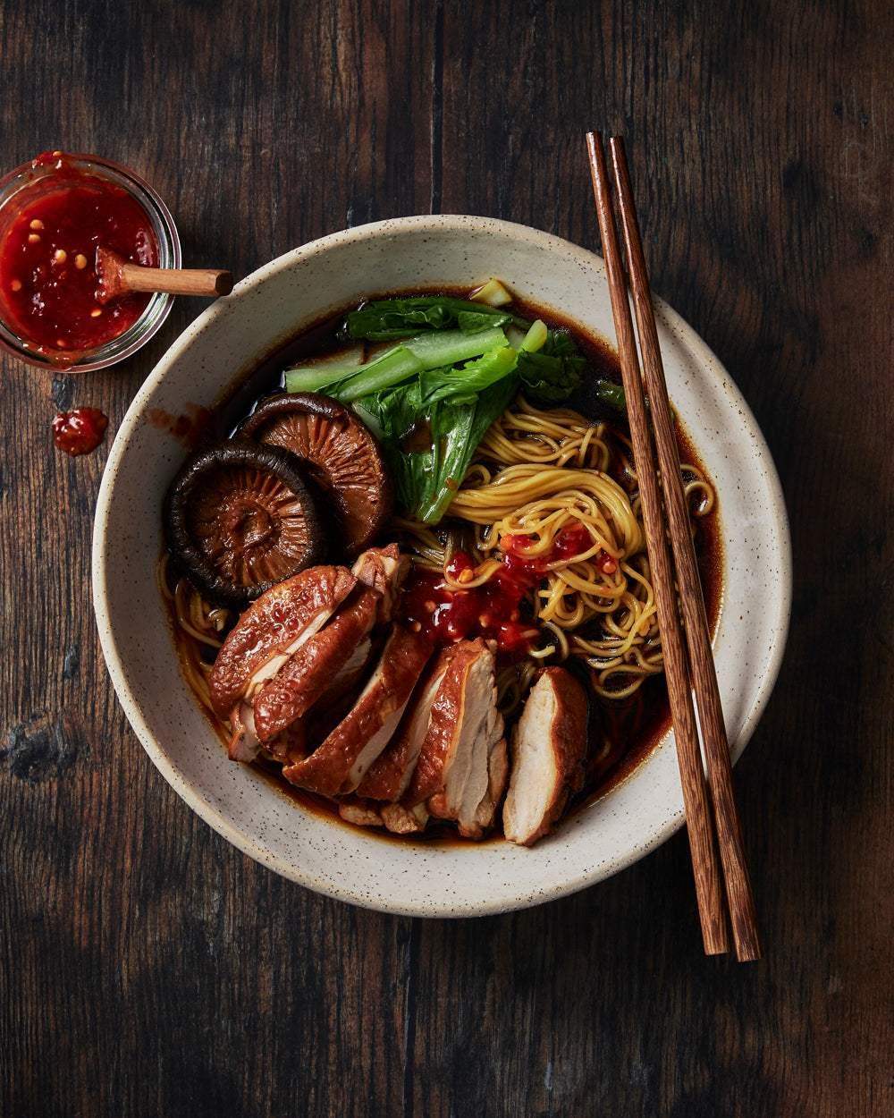 Bowl of Asian noodle soup with sliced chicken, shiitake mushrooms, greens, chili sauce, and chopsticks on wooden table