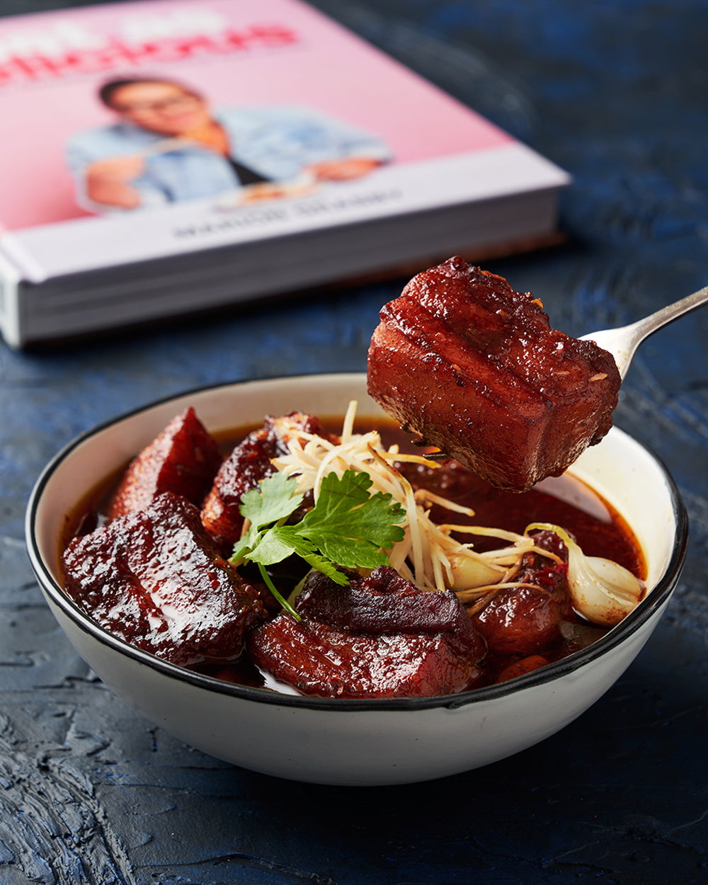Bowl of glazed braised pork with garnish and cookbook in background on blue surface