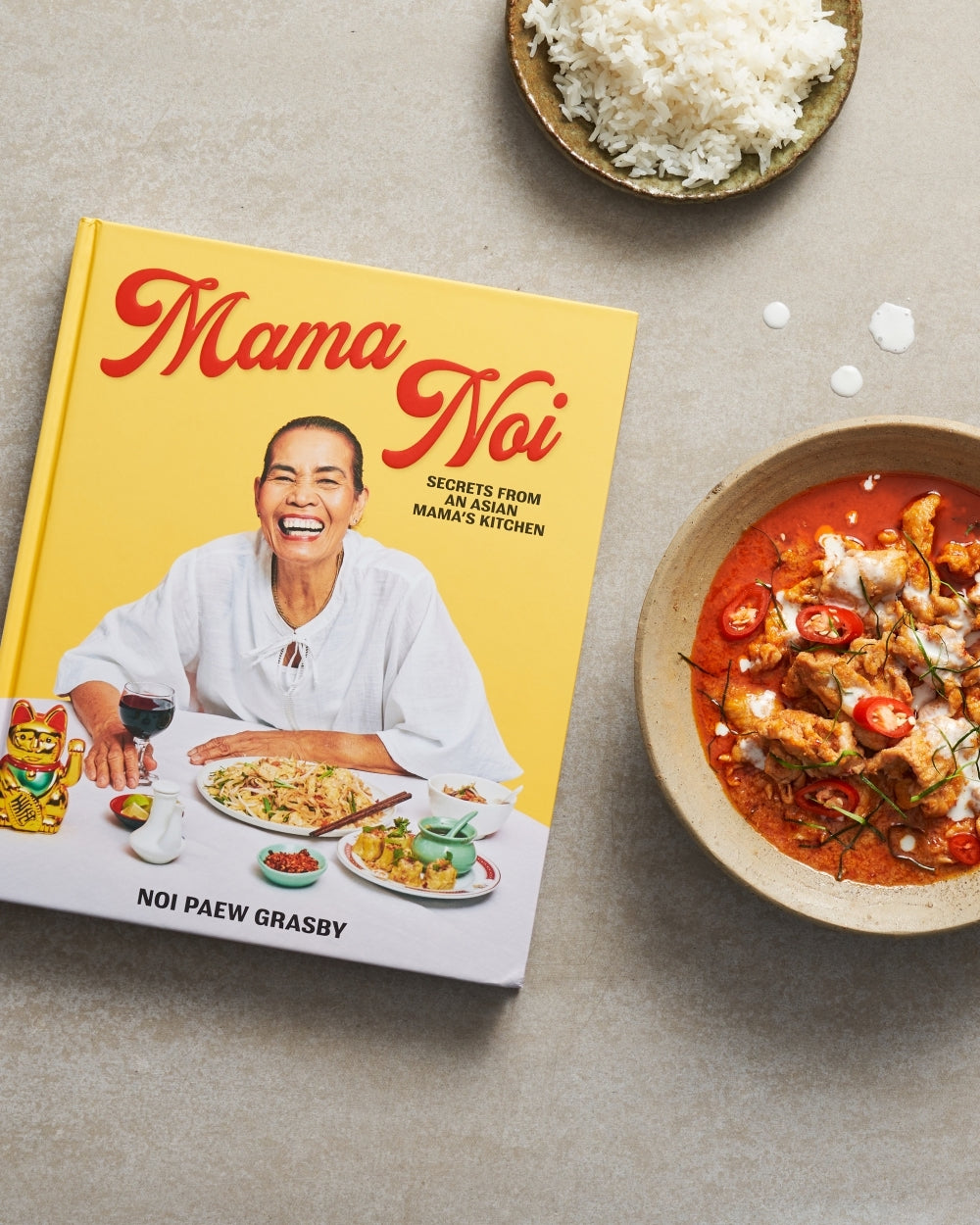 Mama Noi Asian cookbook beside bowl of white rice and spicy curry on a gray table