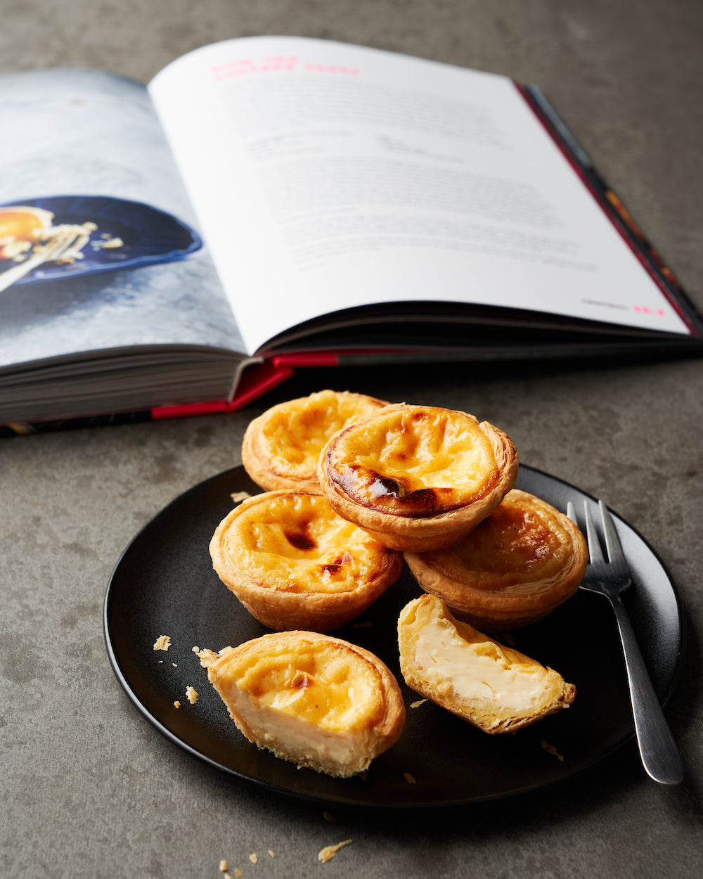 Plate of custard tarts with a fork beside an open cookbook on a grey countertop
