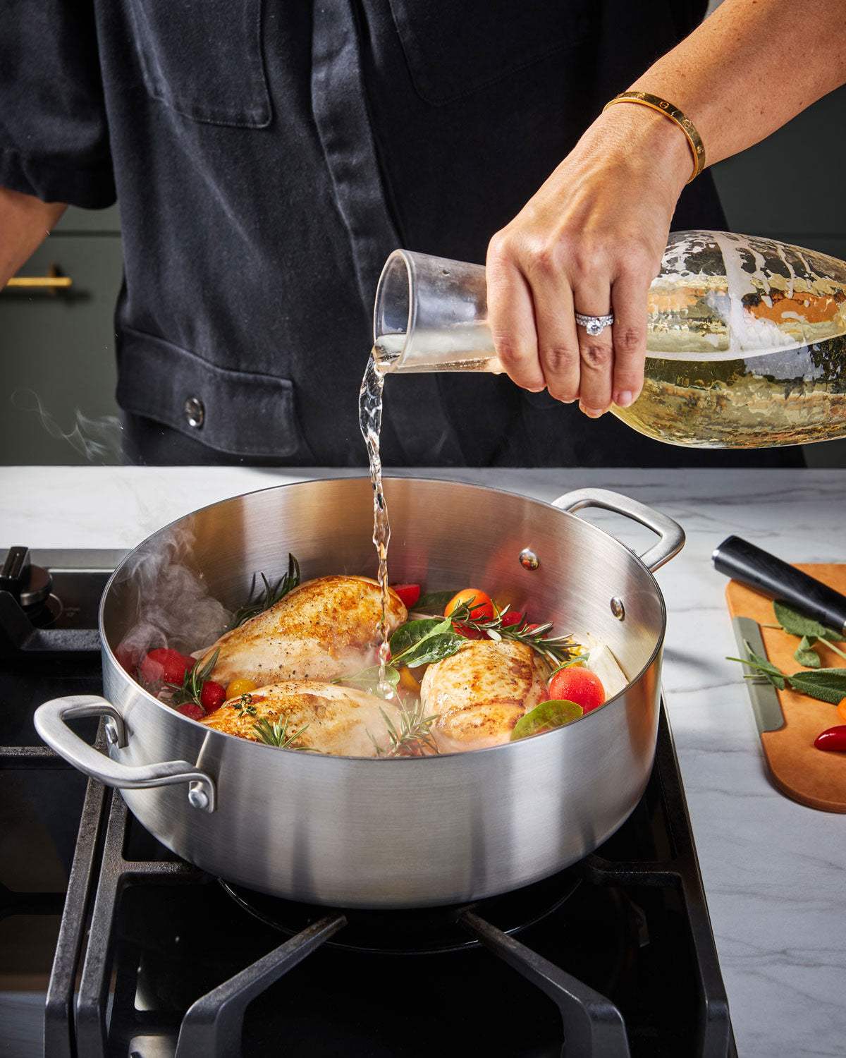 Person pouring liquid into stainless steel sauté pan with chicken breasts and herbs on stovetop
