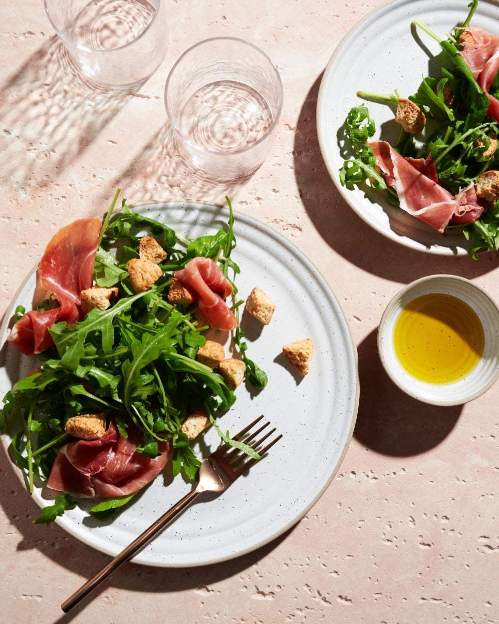 Prosciutto and arugula salad with croutons on white plates, olive oil dip, and water glasses