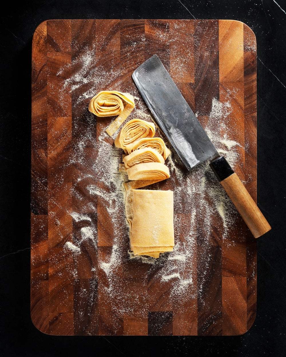 Wooden chopping board with pasta dough, flour, and a cleaver knife on dark countertop