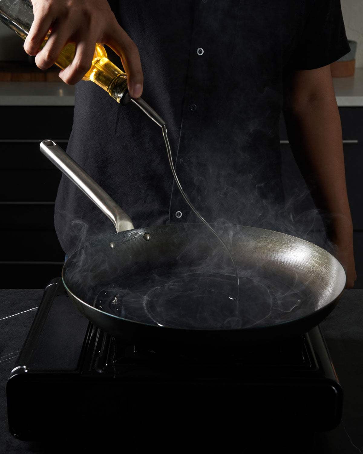 Pouring oil into a hot skillet on stovetop, kitchen cookware in use with visible steam.