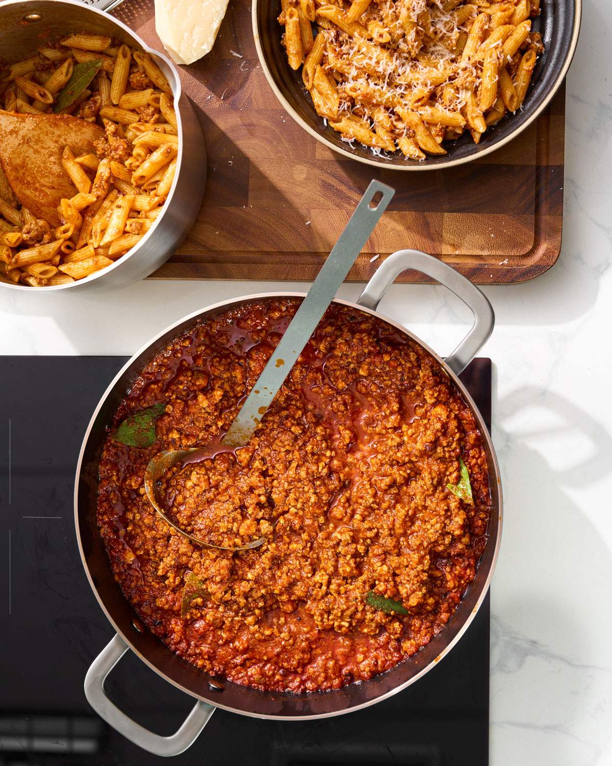 Bolognese sauce simmering in a pan beside cooked penne pasta and grated cheese.