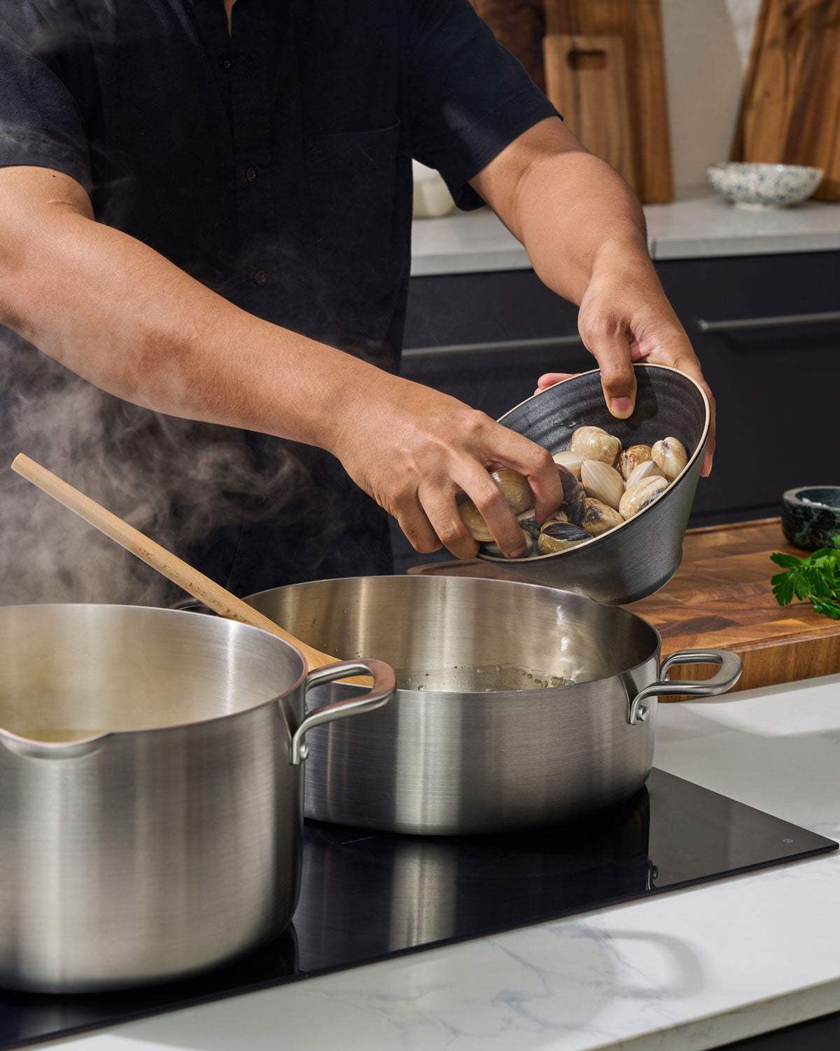 Person adding fresh clams to stainless steel pot on stovetop in modern kitchen