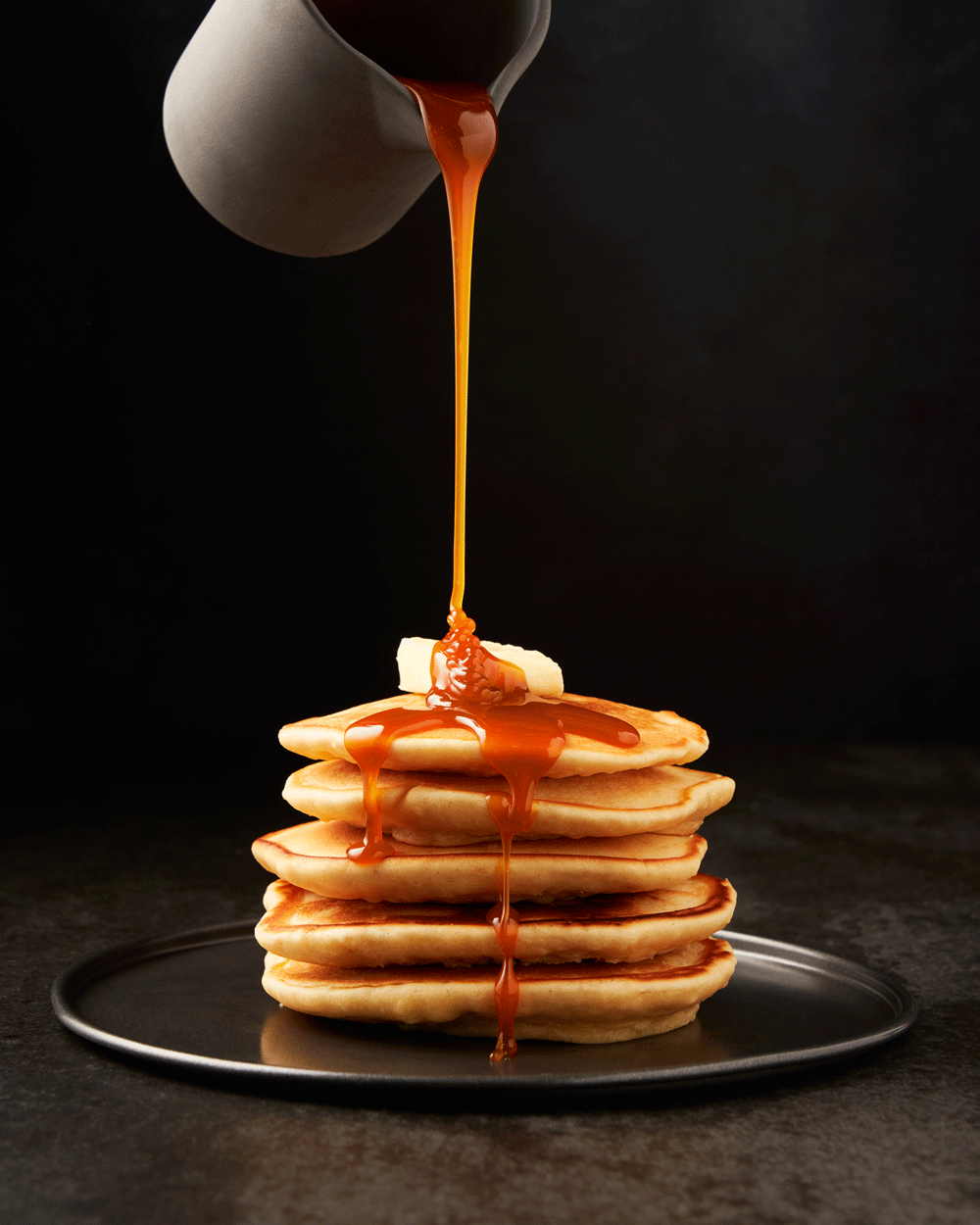 Stack of golden coconut pancakes on dark plate with butter and salted caramel sauce pouring
