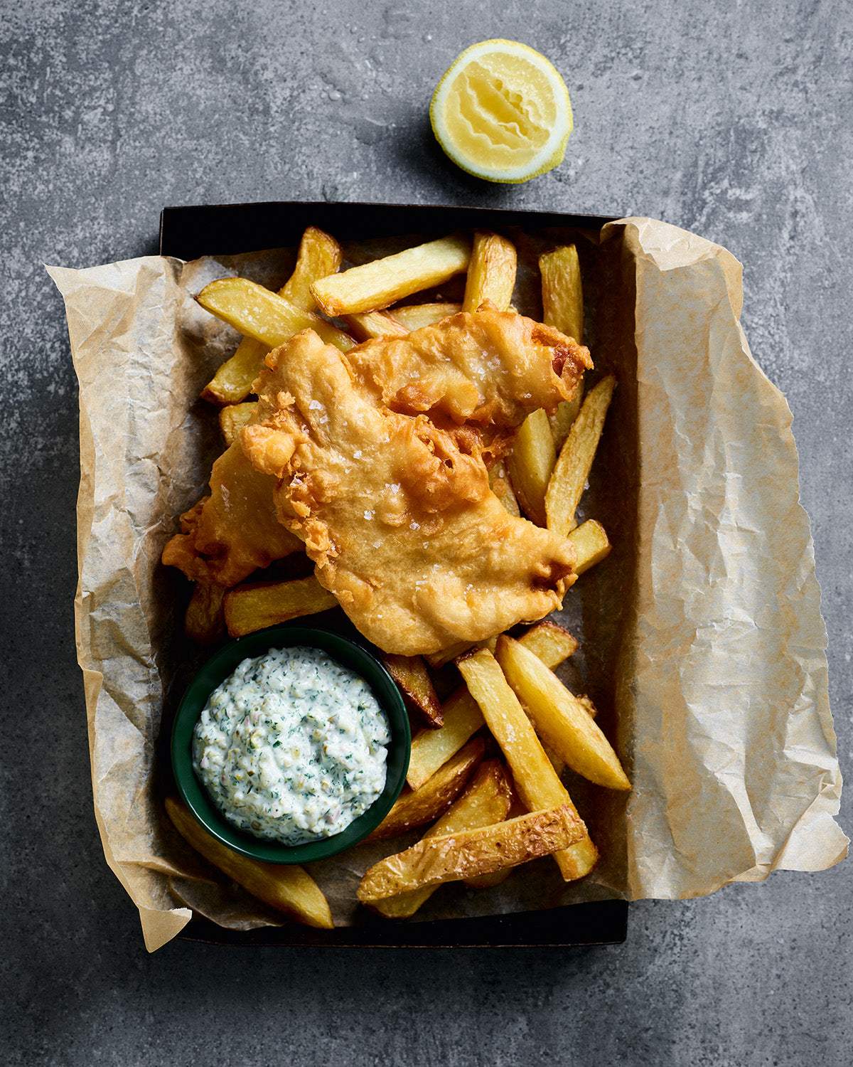 Crispy fish and golden chips with tartar sauce and lemon wedge on parchment paper