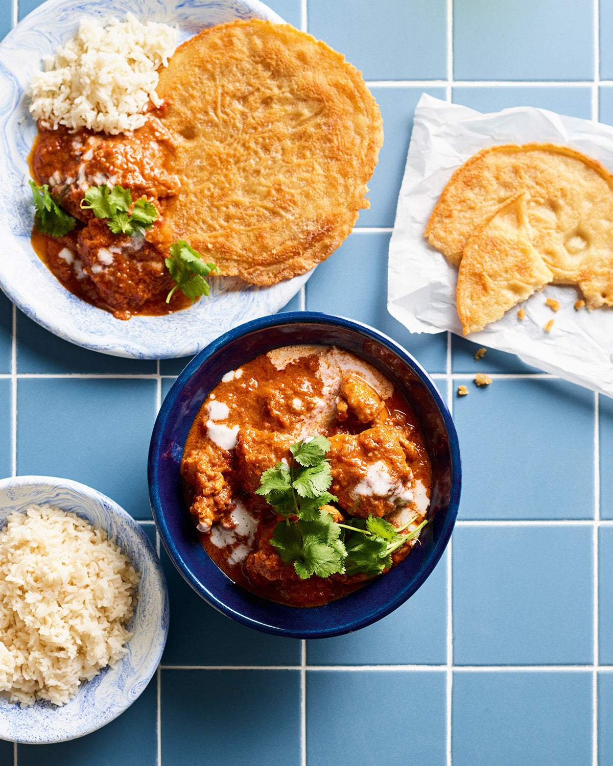 Butter chicken curry garnished with coriander in blue bowl with rice and flatbread on blue tiled table