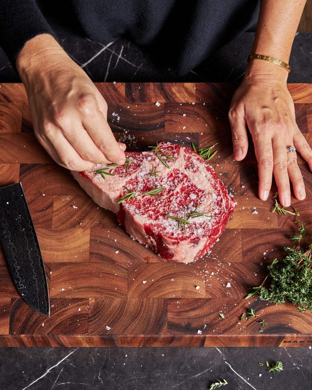 Seasoning raw steak with salt and rosemary on a wooden cutting board beside a chef knife