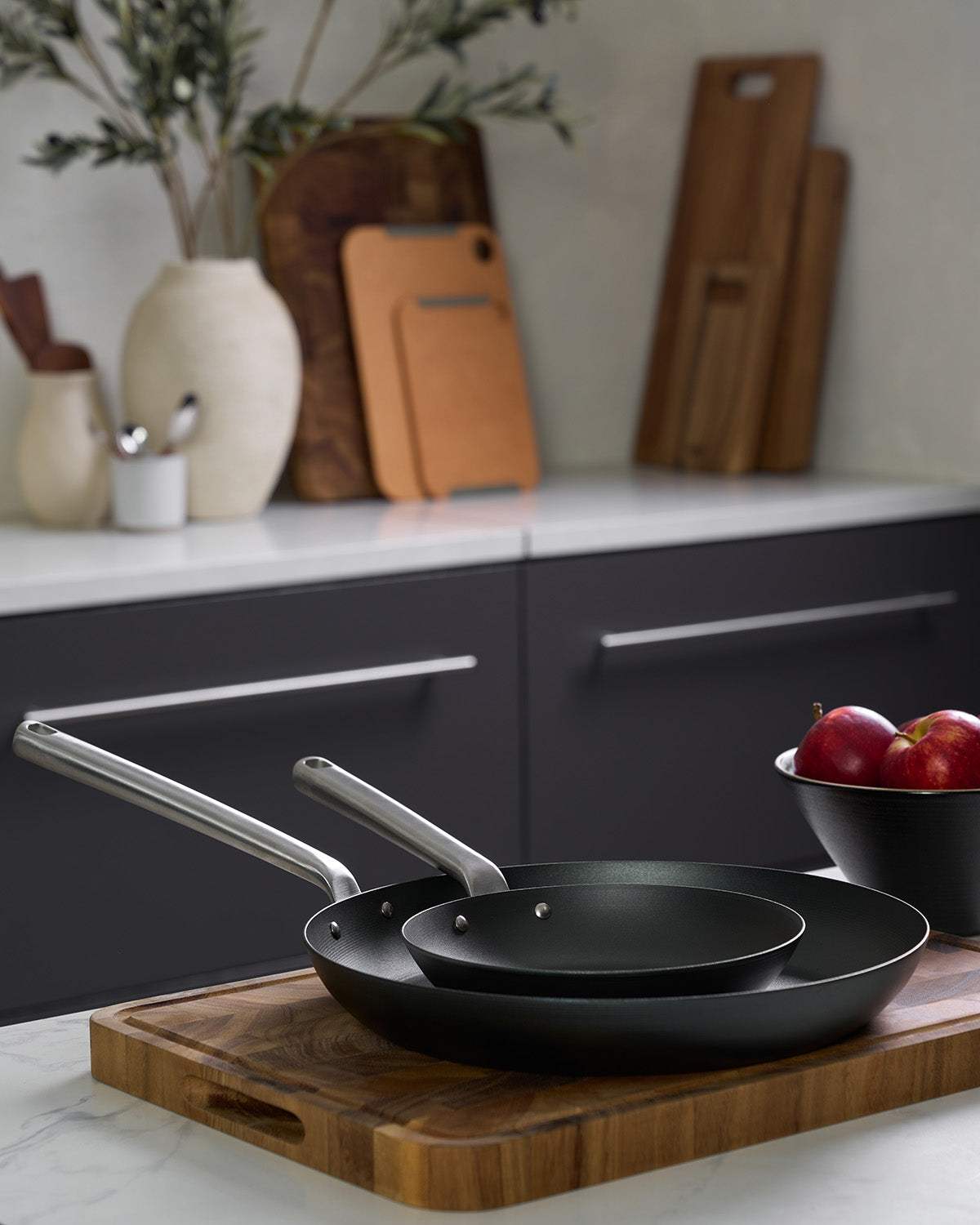 Two black Mako steel frying pans with stainless steel handles on a wooden board in modern kitchen