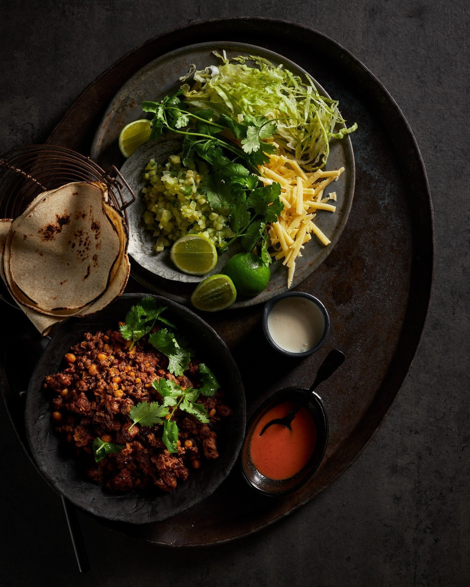 Tex Mex beef taco ingredients with tortillas, seasoned beef, shredded lettuce, cheese, lime, cilantro, and sauces on dark platter