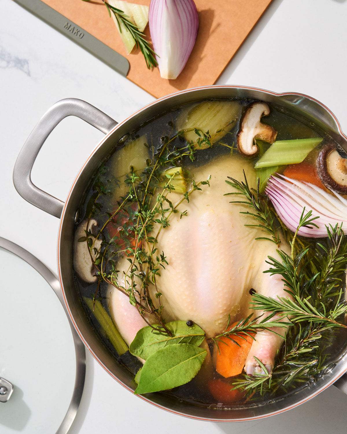 Whole chicken simmering with rosemary, thyme, bay leaves, carrots, celery, and red onion in a Mako pot