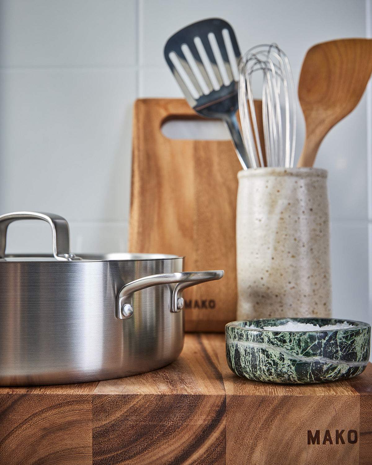 Stainless steel pot, wooden cutting board branded Mako, ceramic utensil holder with kitchen tools, and marble salt cellar on wood block