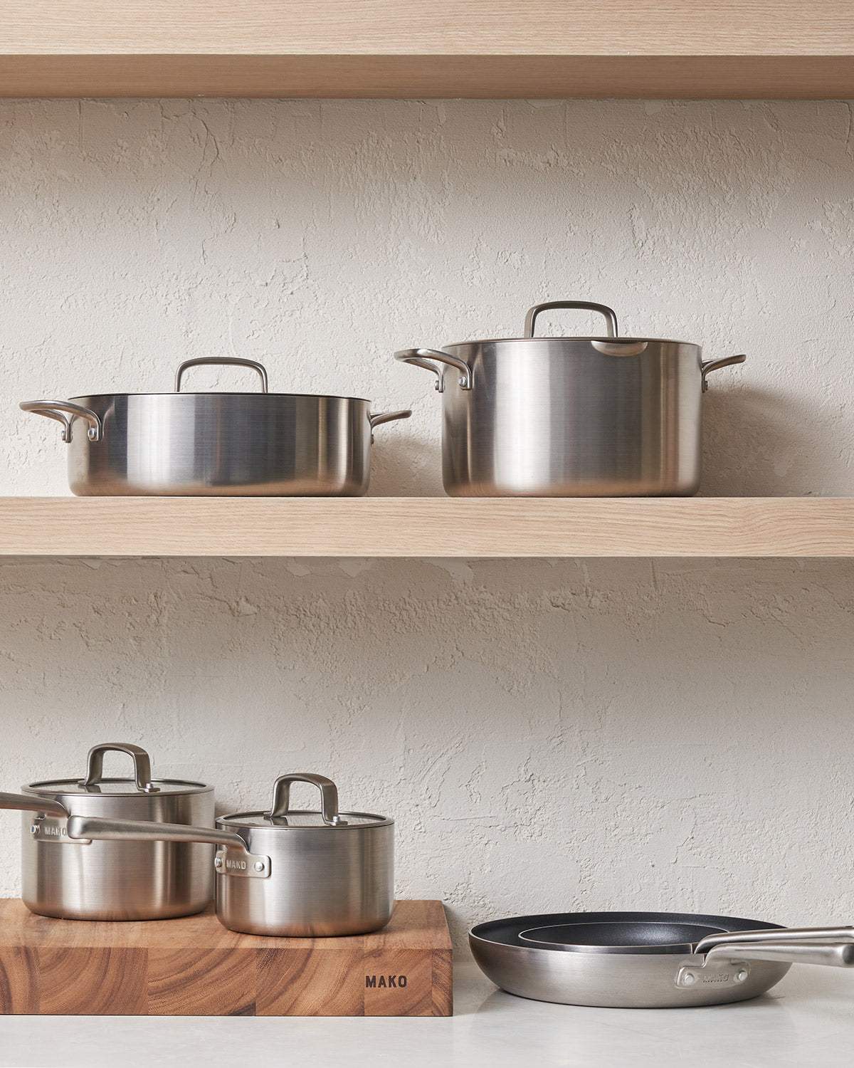 Stainless steel cookware set with pots and frying pans on wooden shelves in a modern kitchen