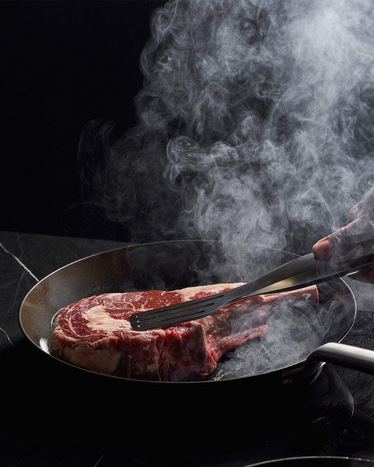 Searing a raw ribeye steak in a black nonstick skillet with metal tongs and rising smoke