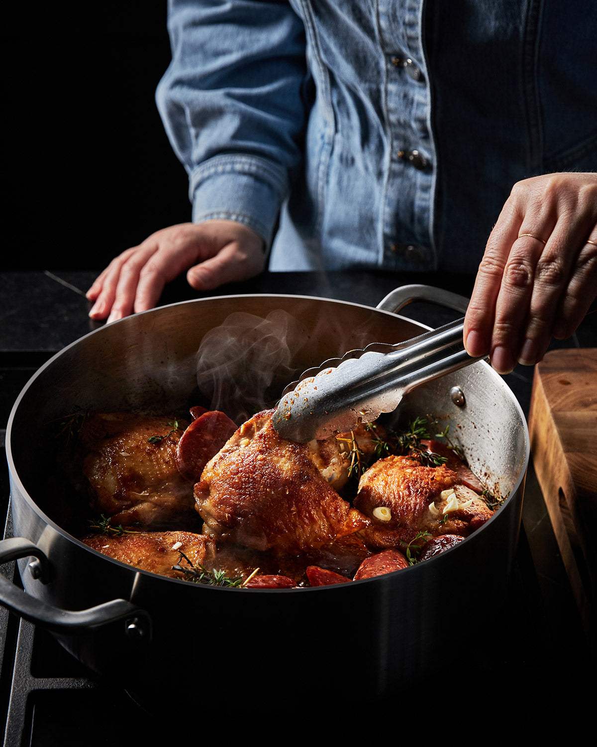 Person cooking crispy chicken thighs with herbs and sausage in a large stainless steel pot