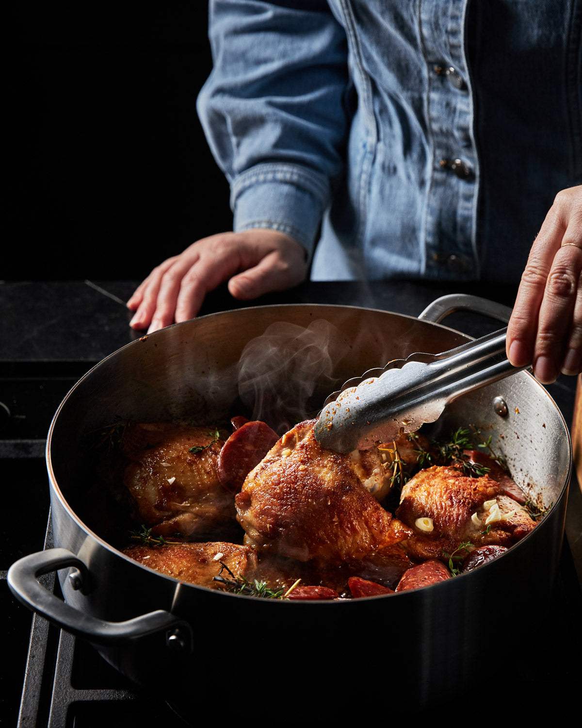 Person cooking crispy chicken thighs with herbs and sausage in a stainless steel pot on stovetop.