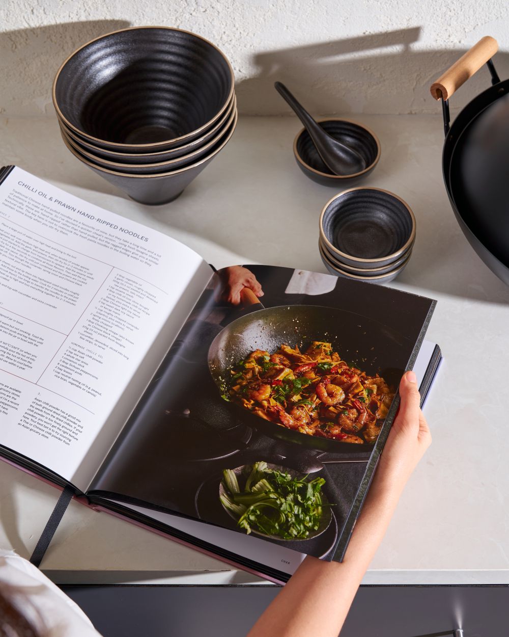 Open cookbook with chili oil prawn noodles recipe, black ceramic bowls, and wok on counter