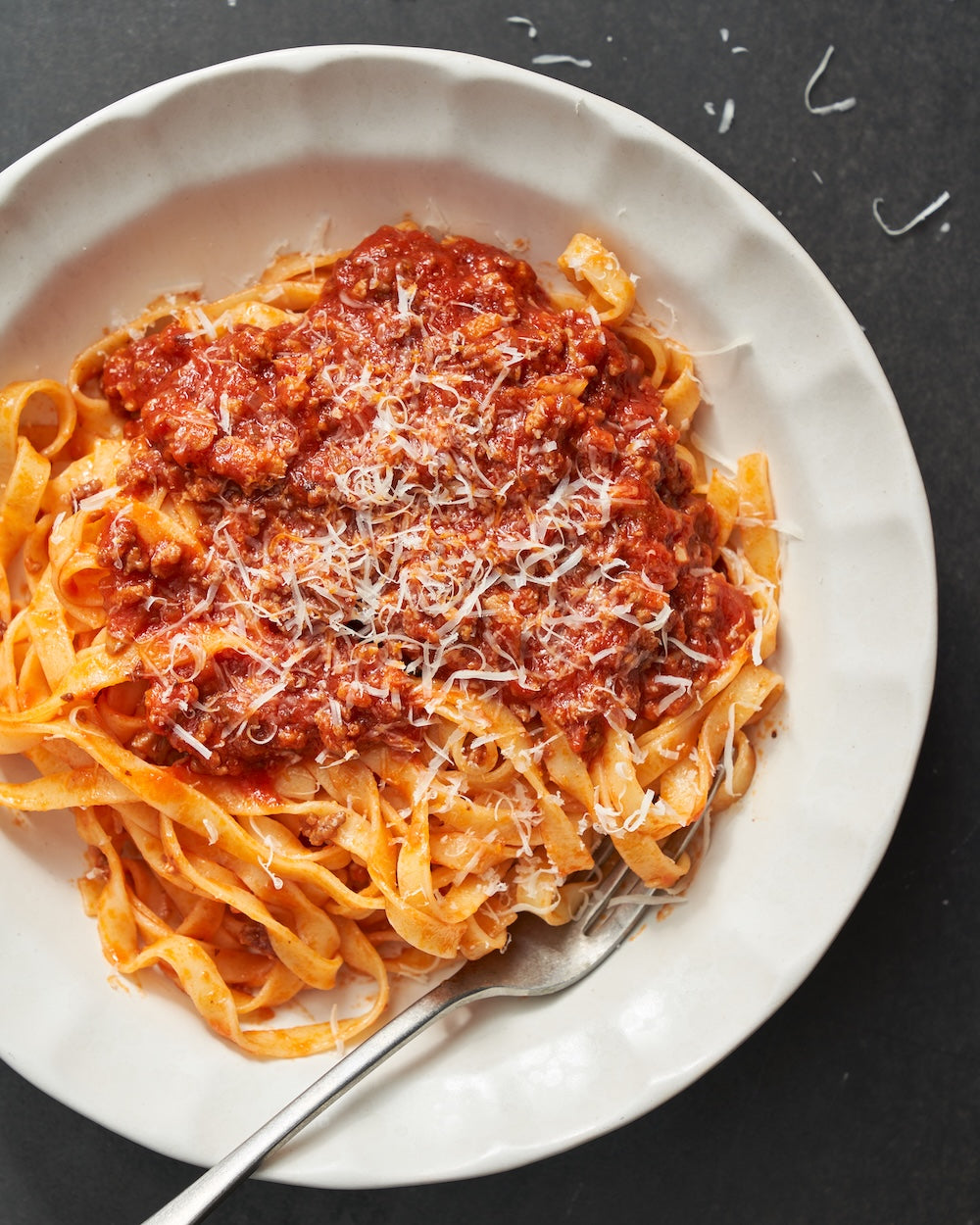 White ceramic bowl of tagliatelle pasta with meat sauce and grated cheese on dark table