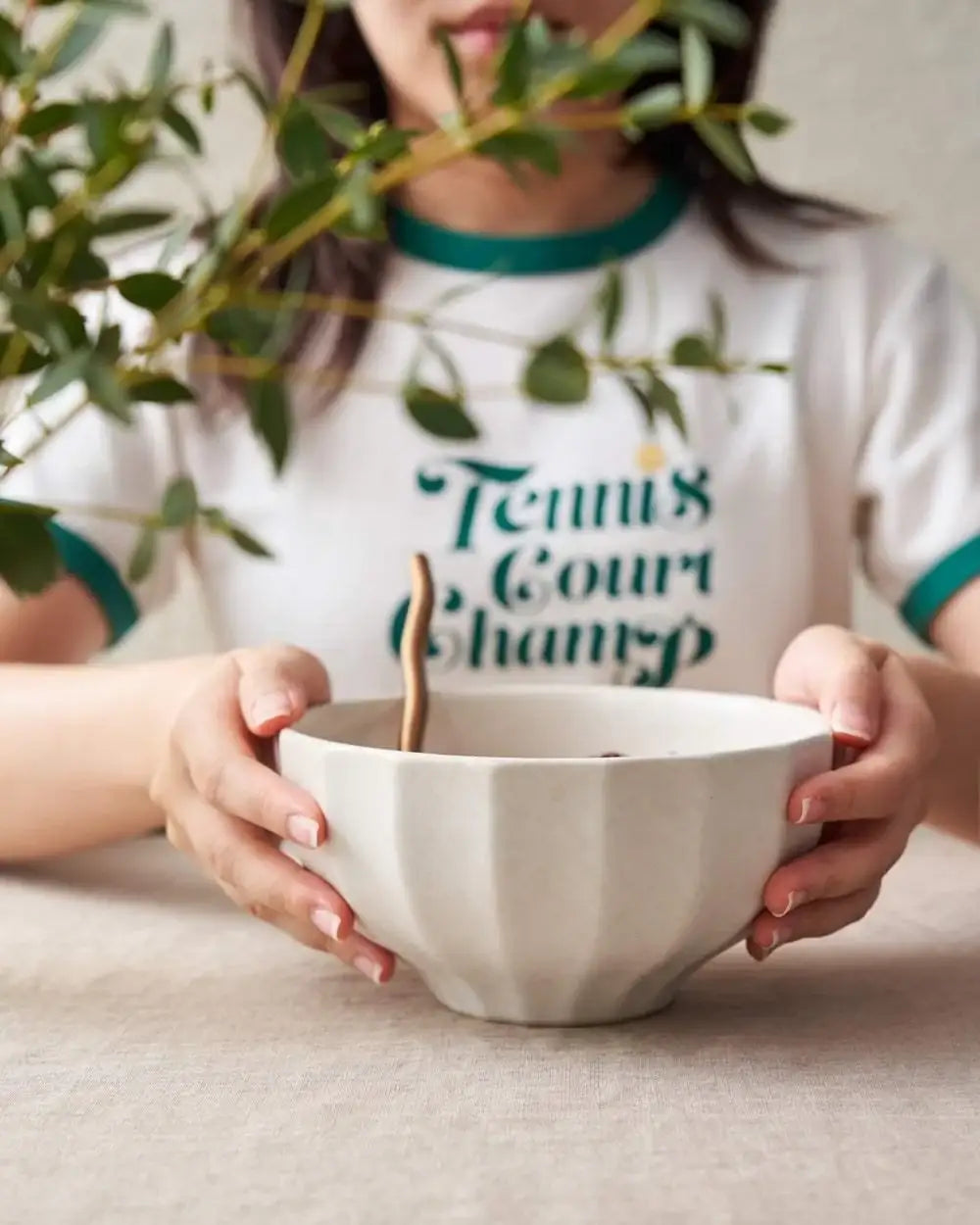 Hands holding white ceramic bowl with wooden spoon, green leaves in foreground, casual setting