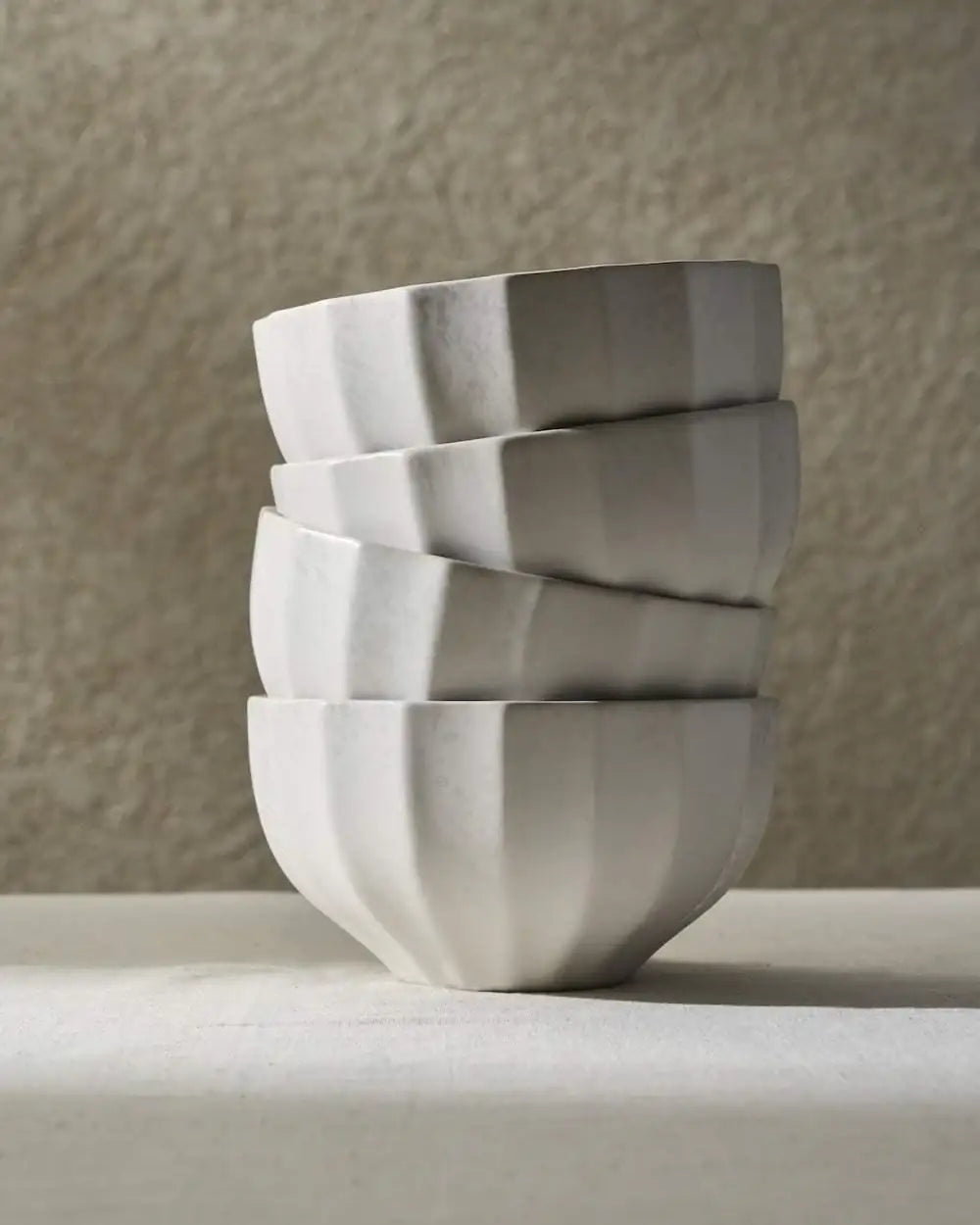 Stack of four white fluted ceramic bowls with soft shadows on a textured beige background