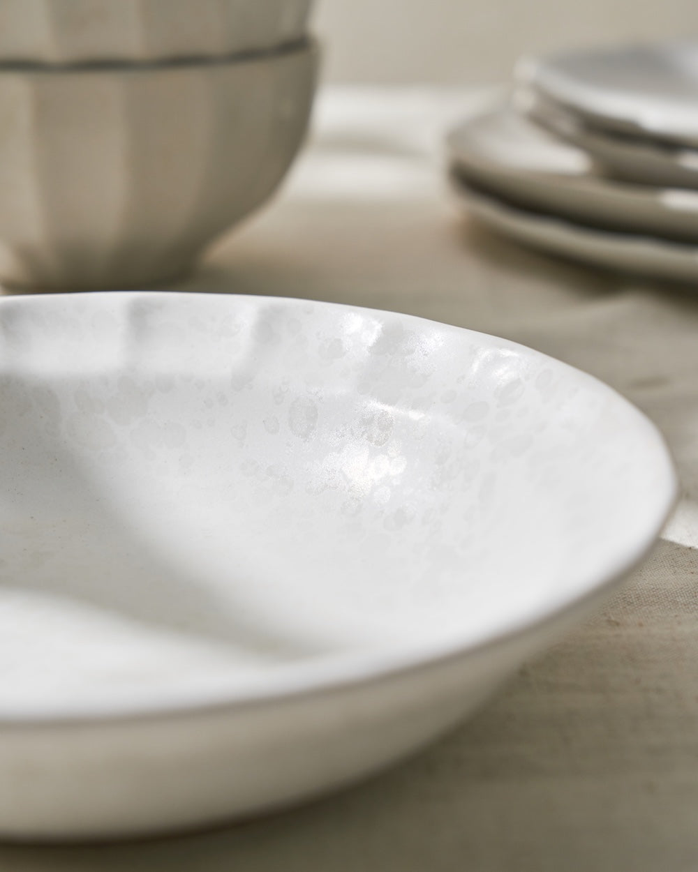 Close-up of handcrafted white ceramic bowl on neutral table with stacked plates in background