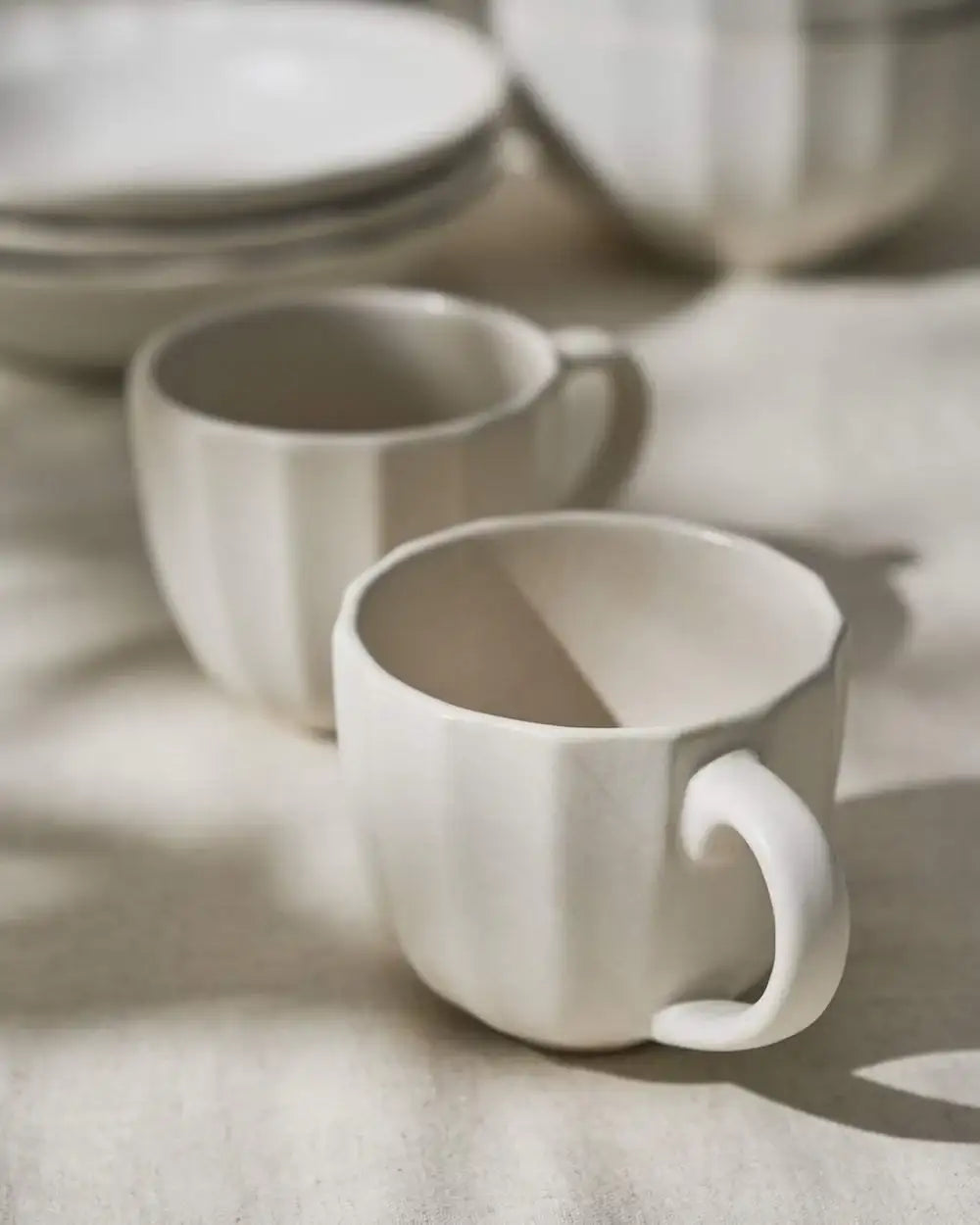 Two cream-colored fluted ceramic mugs on a light fabric surface in soft morning light