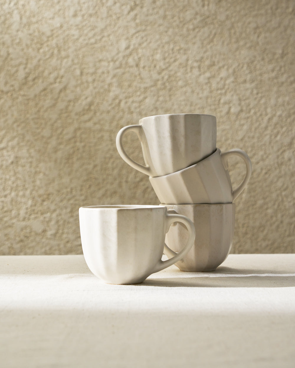 Stacked neutral ceramic mugs with handles on textured beige table and wall