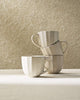 Stack of four cream-colored ceramic mugs with subtle ribbed texture on a light surface, neutral background