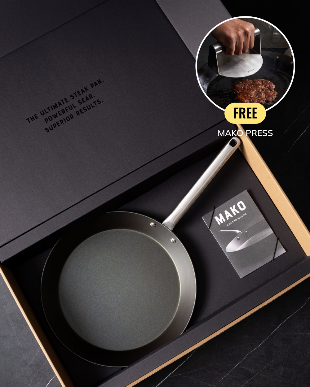 A matte black Mako steak pan with a silver handle presented in a black gift box, alongside a booklet and a circular inset showing a hand using the pan with a Mako press.