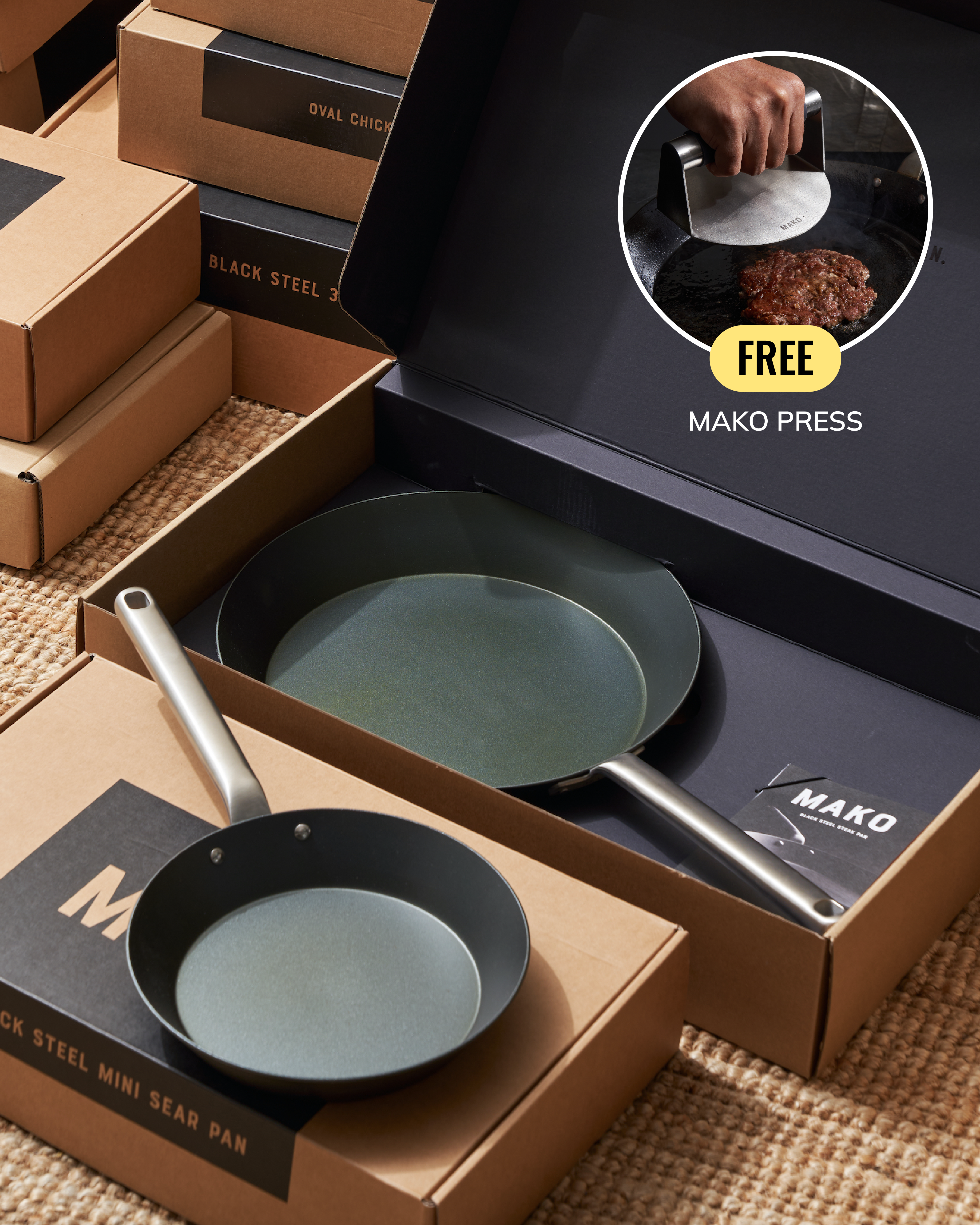 Mako brand cookware gift set featuring black steel mini sear pans in brown cardboard packaging, with an inset circle showing a hand using a Mako press on meat and the text 'FREE MAKO PRESS'.