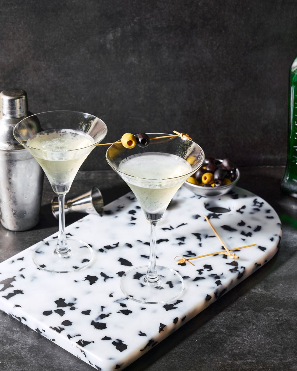 Two martini glasses with olives on a marble serving board, cocktail shaker and olives nearby