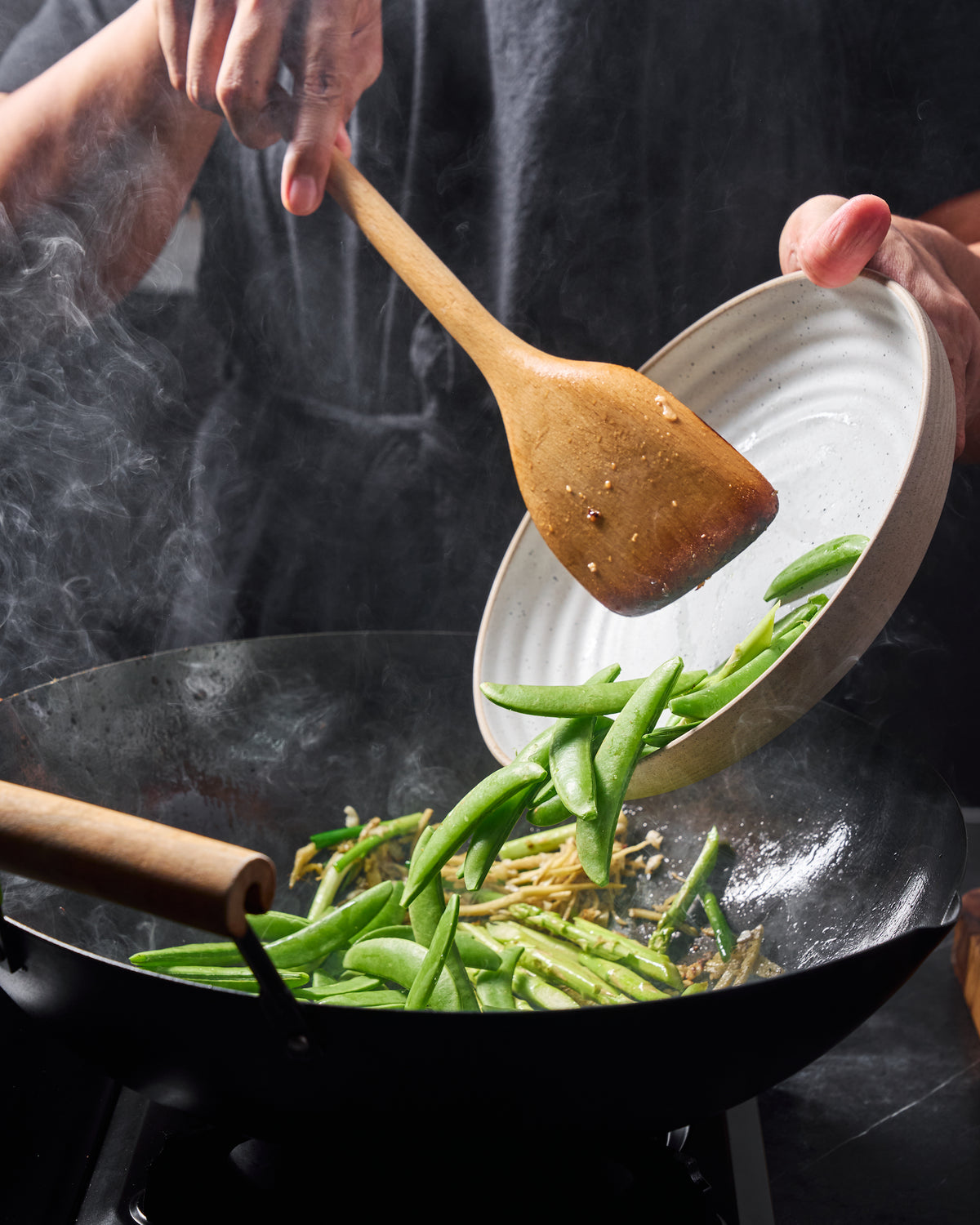 Fresh snow peas being added to a hot black steel wok with asparagus and aromatics during stir-frying.