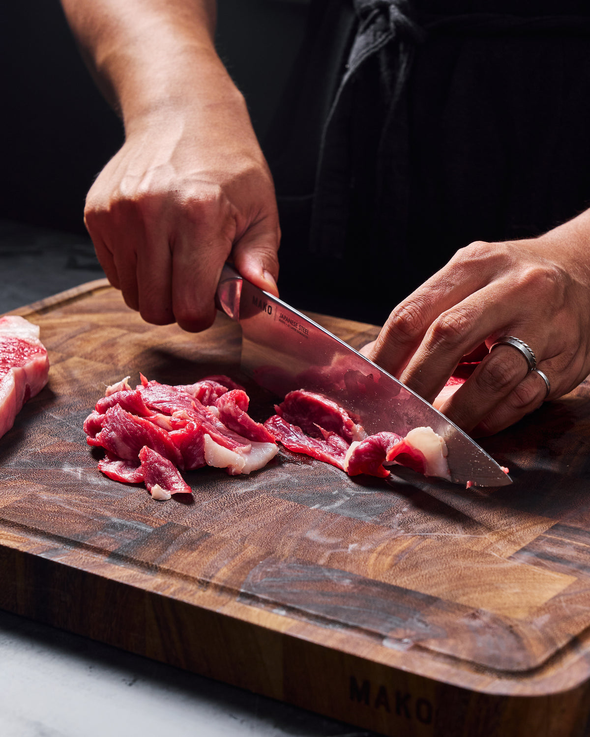 Hands slicing raw beef into thin strips with a MAKO Japanese steel chef knife on a wooden chopping board.