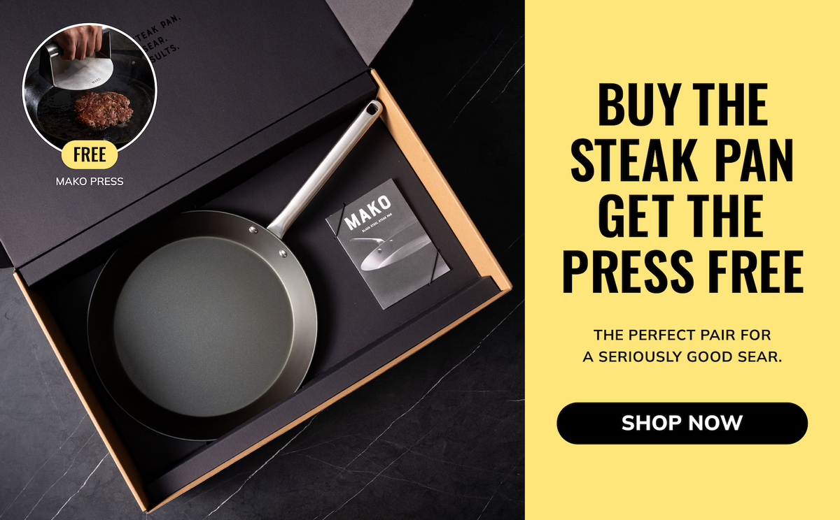 Mako Press steak pan in a black presentation box with a yellow promotional text panel on the right. The pan is dark gray with a silver handle, and the box is shown open on a dark marble surface.