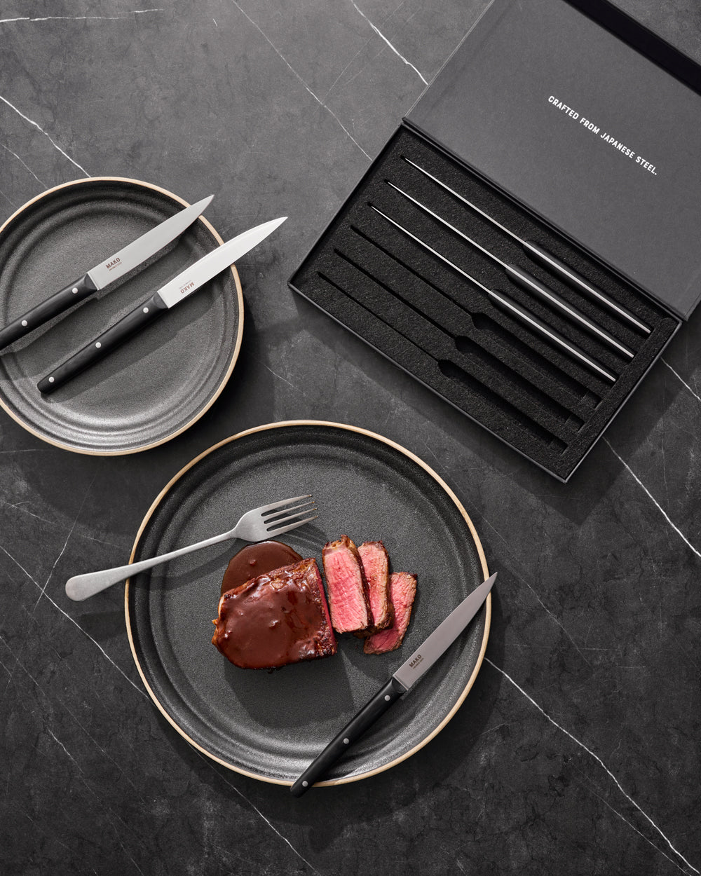 MAKO Steak Knife Set of 6 - Black with steak sliced on charcoal Anaya plate. Black marble benchtop with box and rest of set.