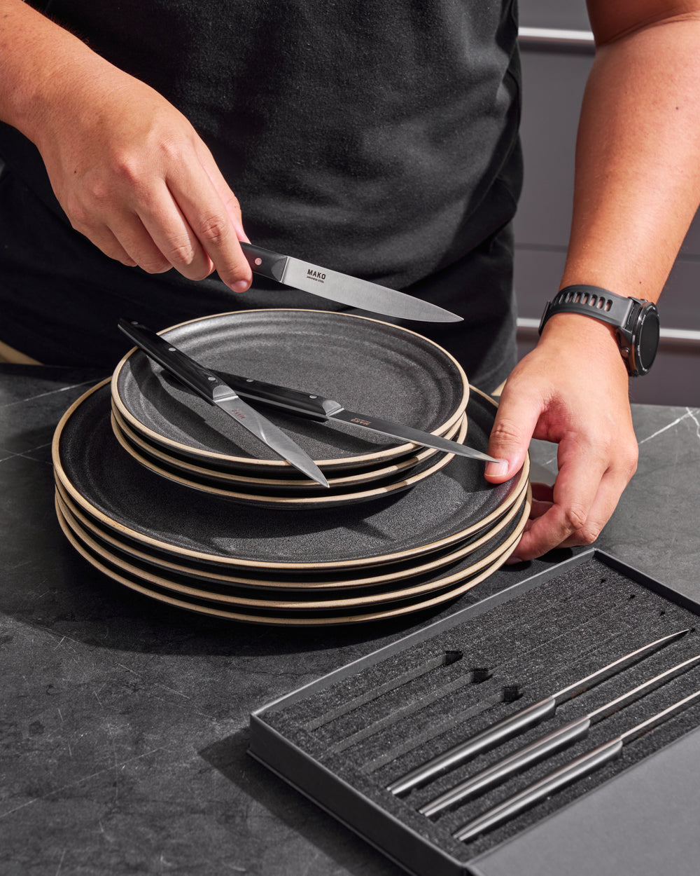 MAKO Steak Knife Set of 6 - Black in hand with rest of set on charcoal Anaya plate stack and in their box.