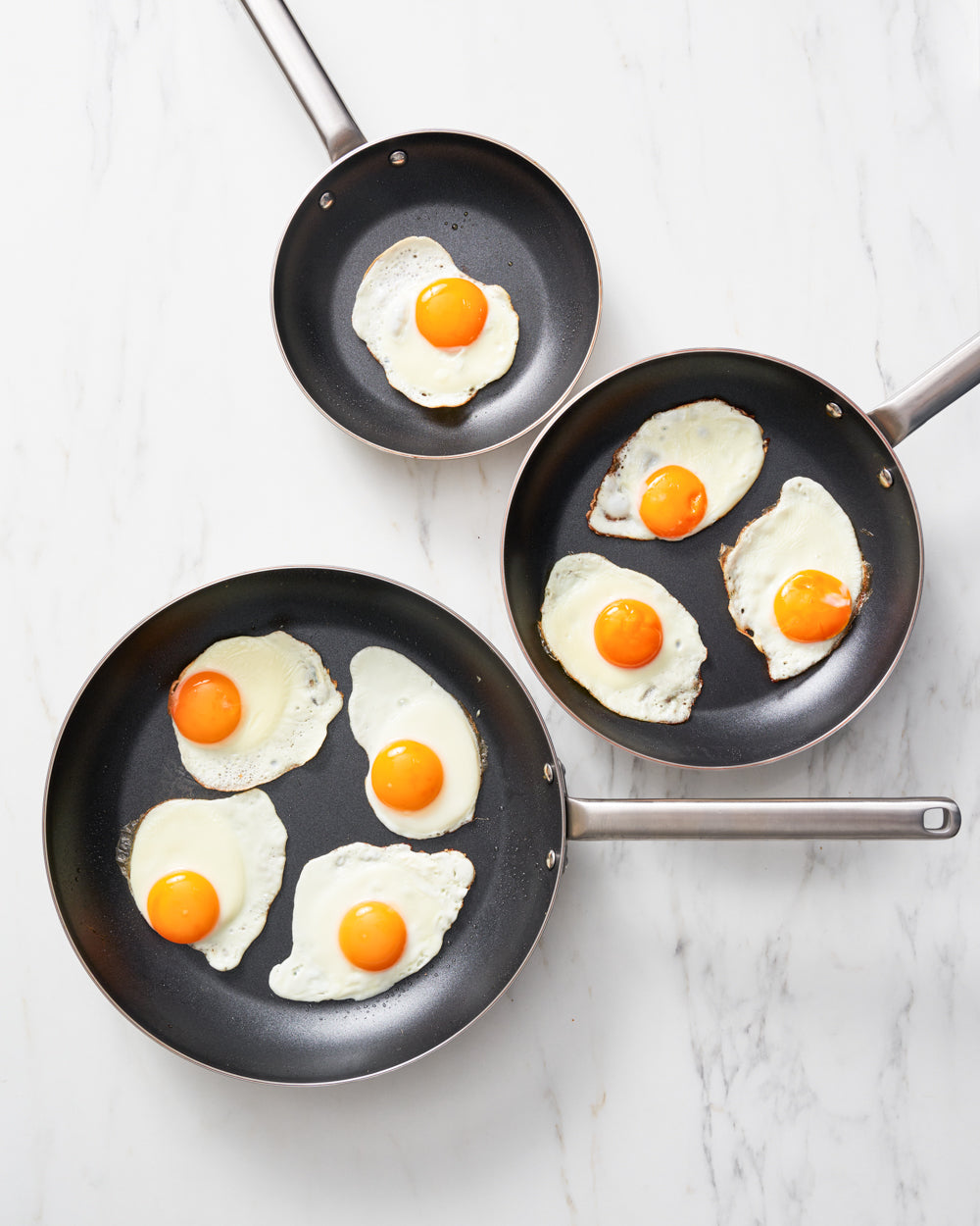 Three non-stick frying pans with sunny side up eggs on a white countertop